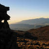 Piton de la Fournaise in the distant early morning light as a hiker descends the summit of Piton des Neiges near cool volcanic rock formations.