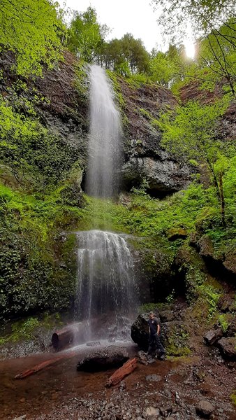 Kilchis Falls drops in a misty veil from a height of perhaps 30 feet, hitting a ledge about halfway down and falling again into a shallow pool, surrounded by greenery.