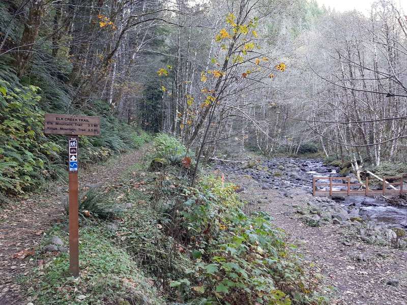 A wooden sign says "Elk Creek Trail" next to a trail that goes along a stream, which has a bridge across it.