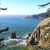 Forested cliffs stretch into the distance with a blue oceans crashing against them below.