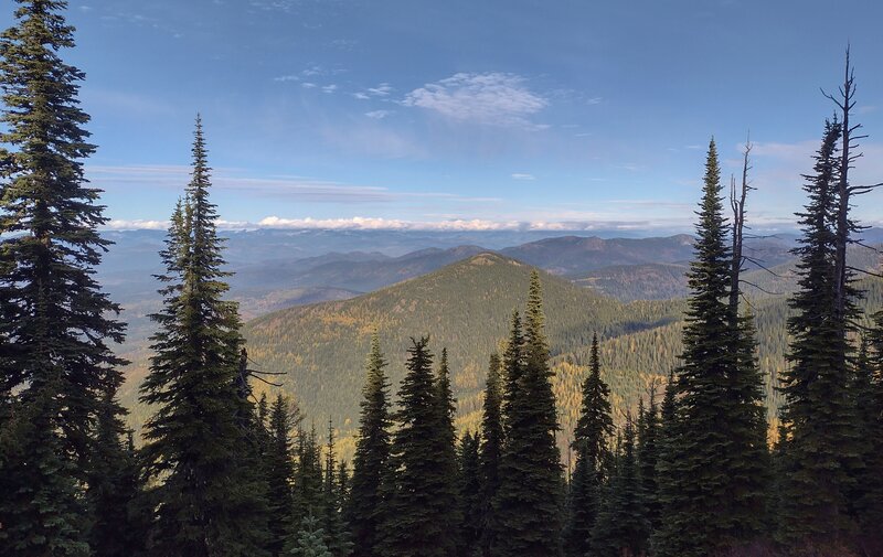 Nearby forested foothills dotted with the bright yellow larches of autumn, are seen below, to the west, from high on the Pend Oreille ridge. Along the horizon, hidden in clouds, in the far distance are Idaho's Selkirk Mountains.